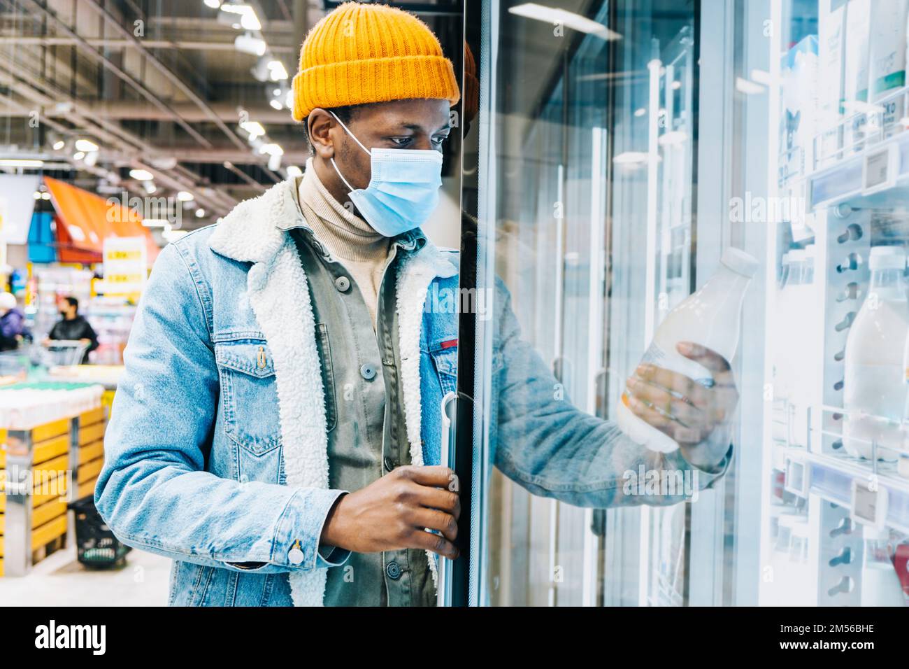 Il giovane uomo afro-americano in giacca denim con maschera usa e getta prende la bottiglia di latte dalla scatola del frigorifero nella moderna sala del supermercato Foto Stock
