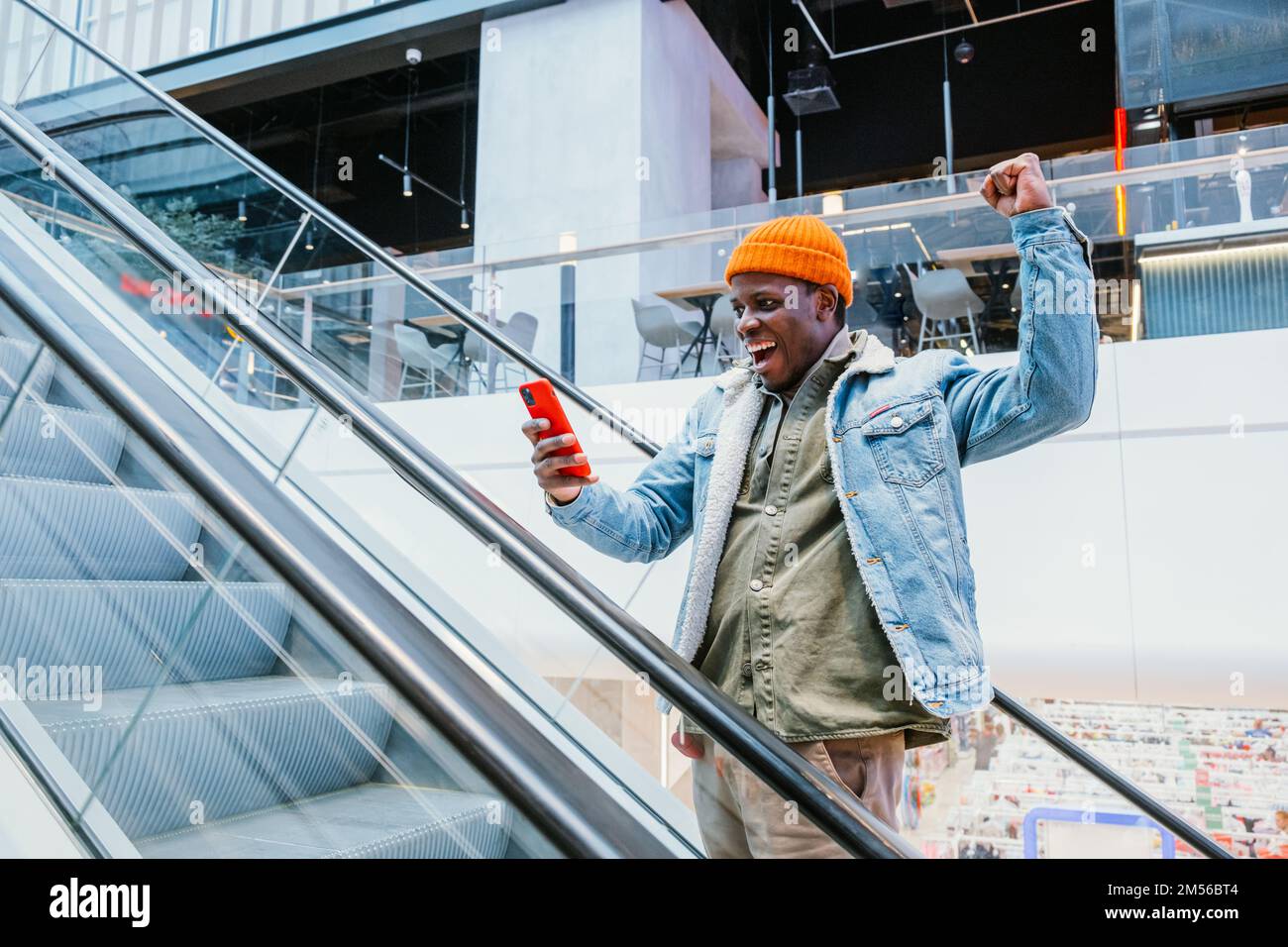 Il giovane afro-americano gioioso in abiti eleganti guarda lo schermo del telefono cellulare che sale scala mobile nel grande centro commerciale Foto Stock
