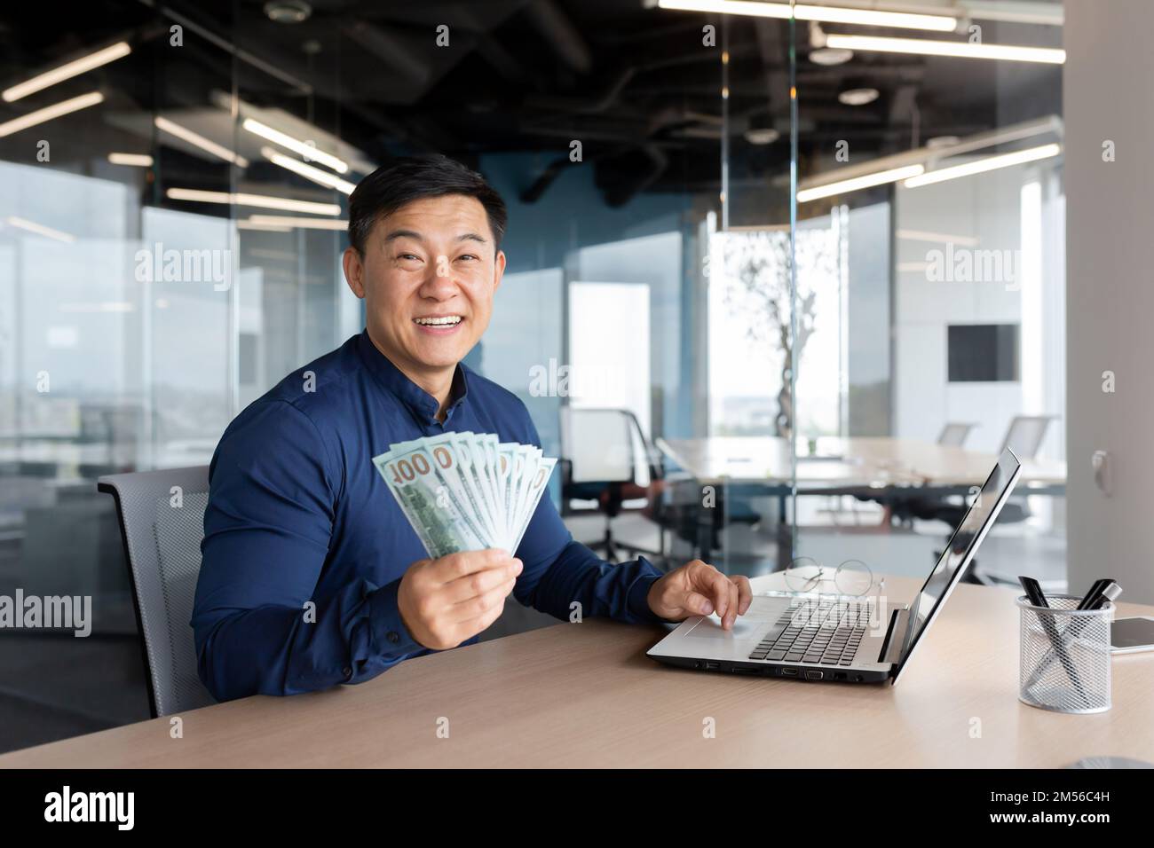 Felice giovane uomo d'affari asiatico seduto in ufficio al tavolo tenendo denaro contante nelle sue mani, mostrando alla fotocamera, sorridendo. Funziona su un computer portatile e documenti. Foto Stock