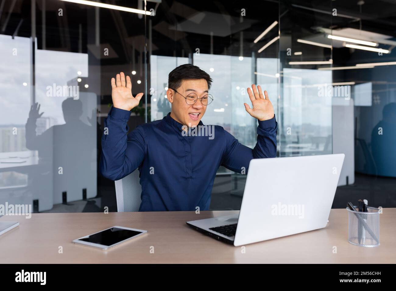 Giovane uomo asiatico libero, uomo d'affari in ufficio al tavolo parlando tramite videochiamata con i partner, incontro online, gioisce, si congratula, ha alzato le mani. Foto Stock