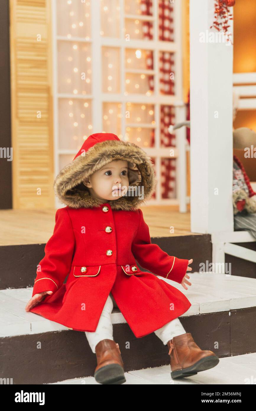 bambino affascinante in un cappotto rosso con un cappuccio siede sul portico Foto Stock