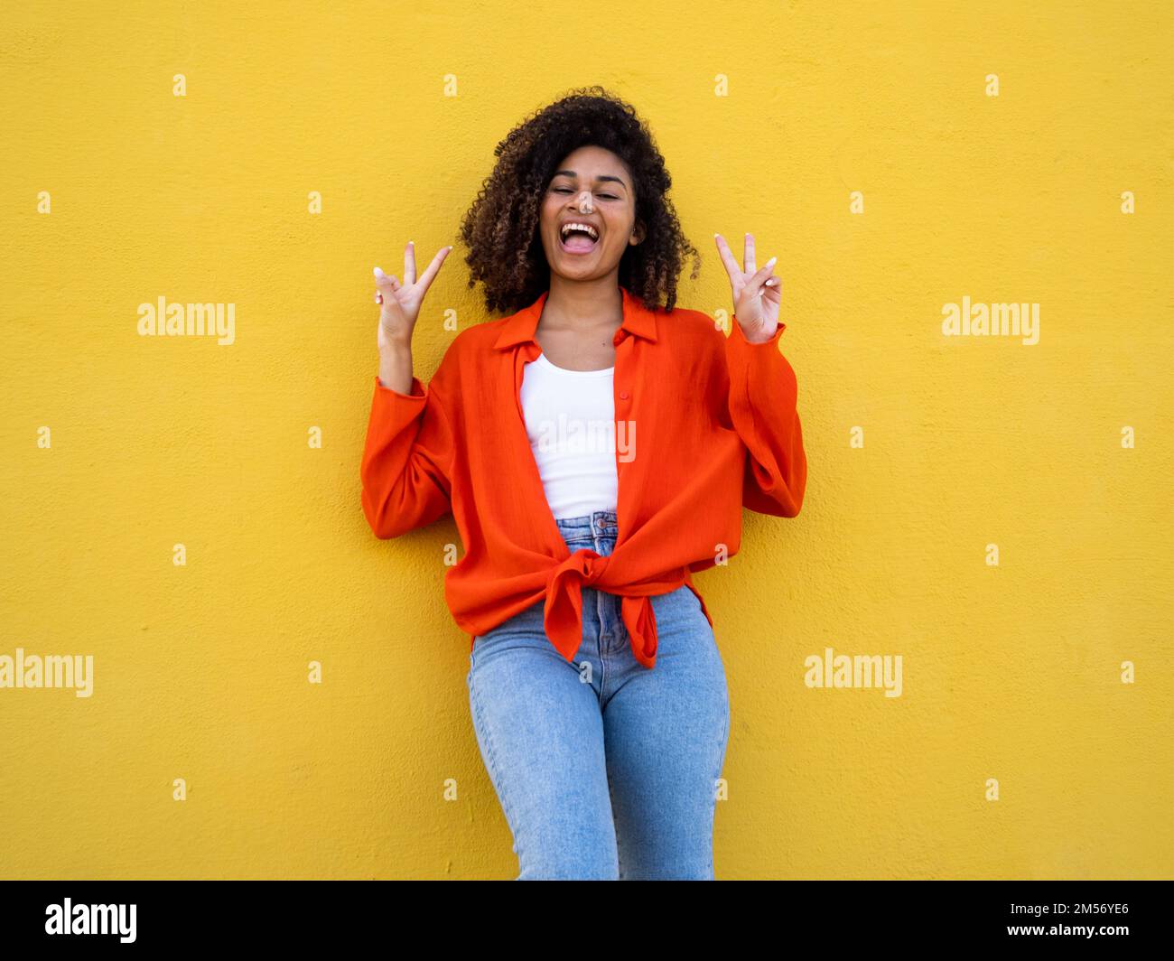 Allegra giovane afroamericana sorridente e simbolo di vittoria con le mani sul muro giallo. Vittoria celebrando il gesto Foto Stock