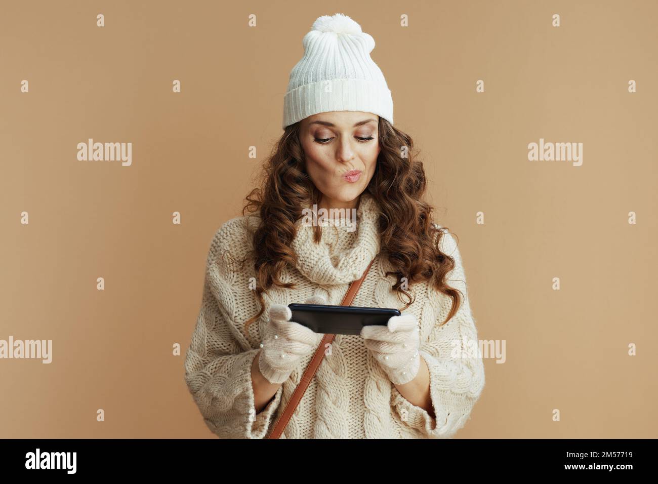 Ciao inverno. donna alla moda in maglione beige, guanti e cappello su sfondo beige utilizzando le applicazioni per smartphone. Foto Stock