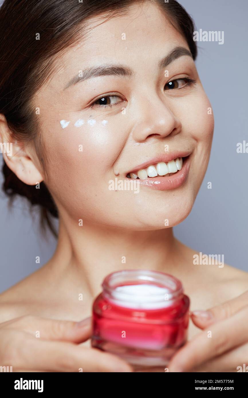 Ritratto di moderna donna asiatica con vasetto crema viso e crema occhi sul viso su sfondo blu. Foto Stock