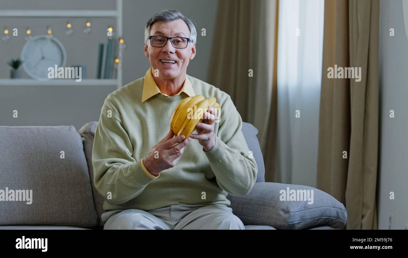 Il vecchio uomo caucasico di 60s anni l'uomo arrabbiato siede sul divano a casa parlando guardando la macchina fotografica blog il gastroenterologo nutrizionista maschio tiene i frutti gialli maturi Foto Stock