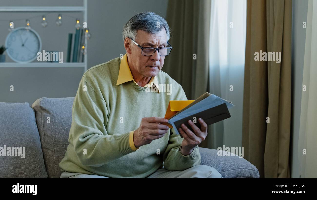 Vecchio uomo caucasico 60s anni che controlla le lettere di smistamento nel soggiorno nonno maturo concentrato seduto sul divano guardando l'avviso di corrispondenza Foto Stock