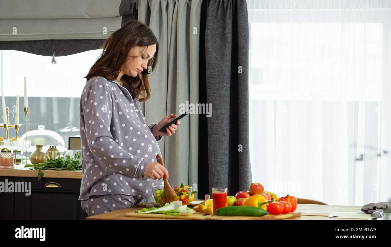 Donna incinta guarda la ricetta al telefono e prepara l'insalata Foto Stock