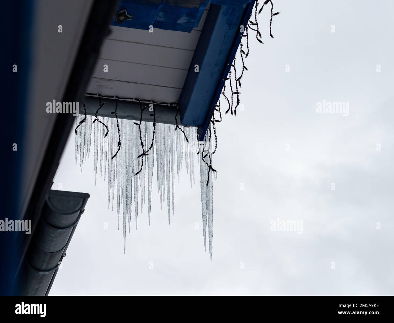 Ghiaccioli appesi su una grondaia del tetto. Le luci decorative si trovano sul tetto in legno. Il cielo è coperto di nuvole. L'acqua di fusione sta gocciolando giù su un edificio. Foto Stock