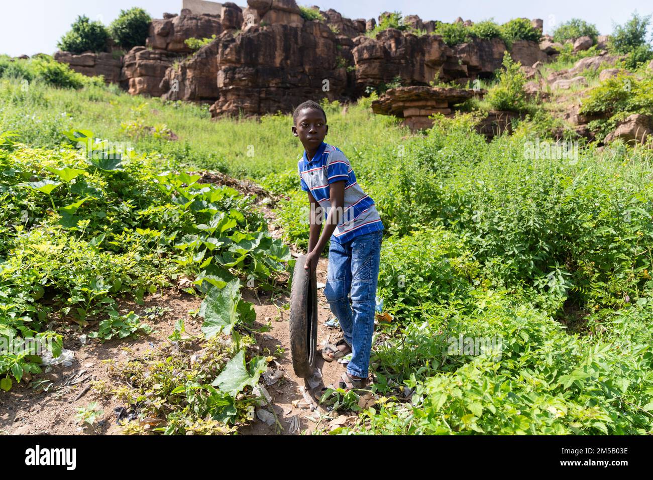 Carino ragazzo africano che gioca da solo con un vecchio pneumatico che lo lascia rotolare giù per la collina su un percorso sterrato Foto Stock