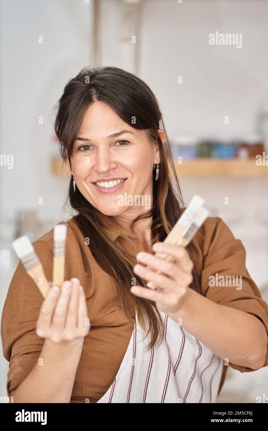 Donna in grembiule che guarda la macchina fotografica e sorridente mentre tiene gli attrezzi di lavoro in un laboratorio di ceramica. Concetto di artigianato, arte e hobby. Foto Stock