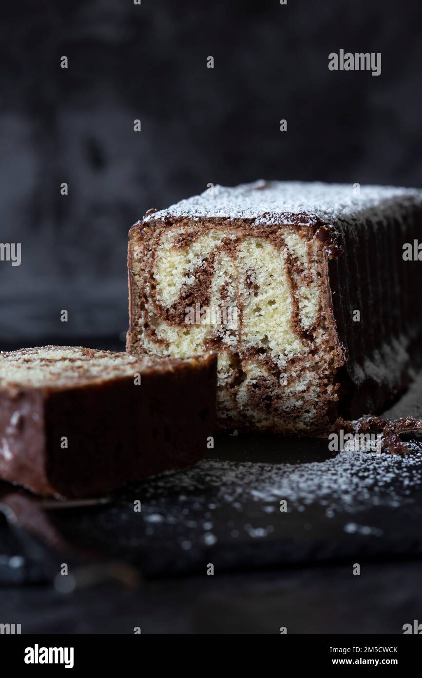 Torta di marmo al cioccolato fatta in casa su un tagliere di ardesia ricoperto di zucchero a velo. Tagliare a pezzi con un affettatore per torte Foto Stock