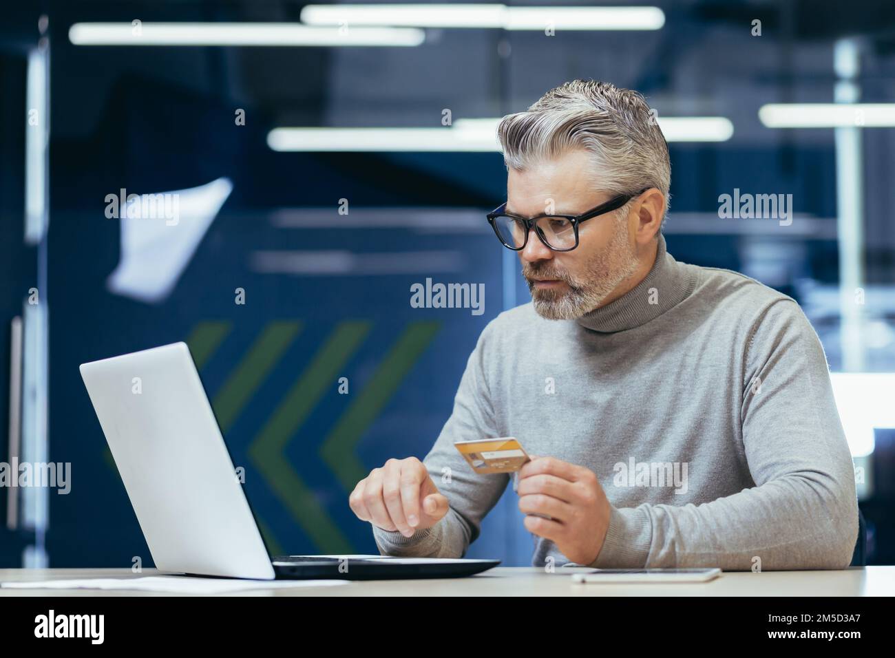 Senior grigio haired uomo d'affari in ufficio frustrato e triste cercando di effettuare la transazione bancaria e l'acquisto in negozio online internet, l'uomo che tiene la carta di credito e utilizzando il computer portatile. Foto Stock