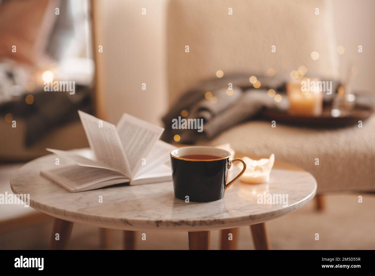 Tazza di tè con libro aperto di carta e candele profumate brucianti sul tavolo di marmo sopra la sedia accogliente e luci scintillanti nel closeup della camera da letto. Vacanze invernali mari Foto Stock