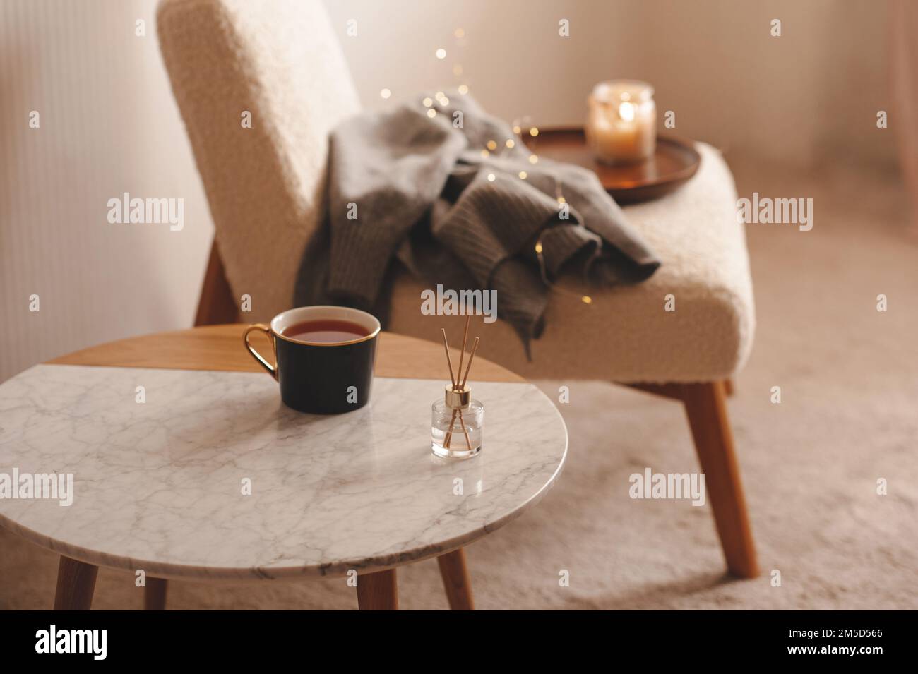 Tazza di tè con diffusore di fragranze liquide su tavolo in marmo sopra sedia con candele e tessuto in maglia in camera da letto closeup. Stagione invernale. Foto Stock