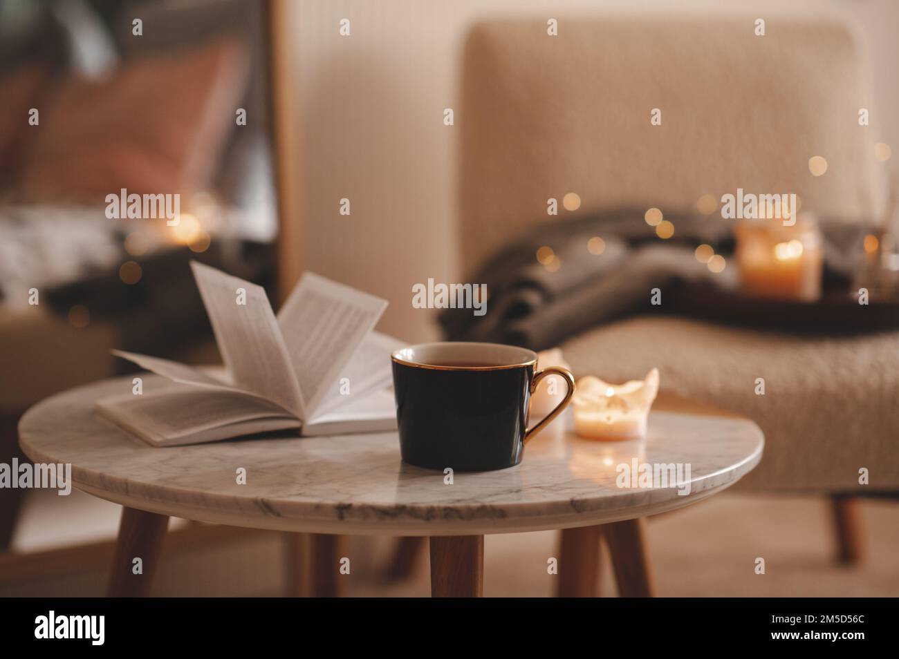Tazza di tè con libro aperto di carta e candele profumate brucianti sul tavolo di marmo sopra la sedia accogliente e luci scintillanti nel closeup della camera da letto. Vacanze invernali mari Foto Stock