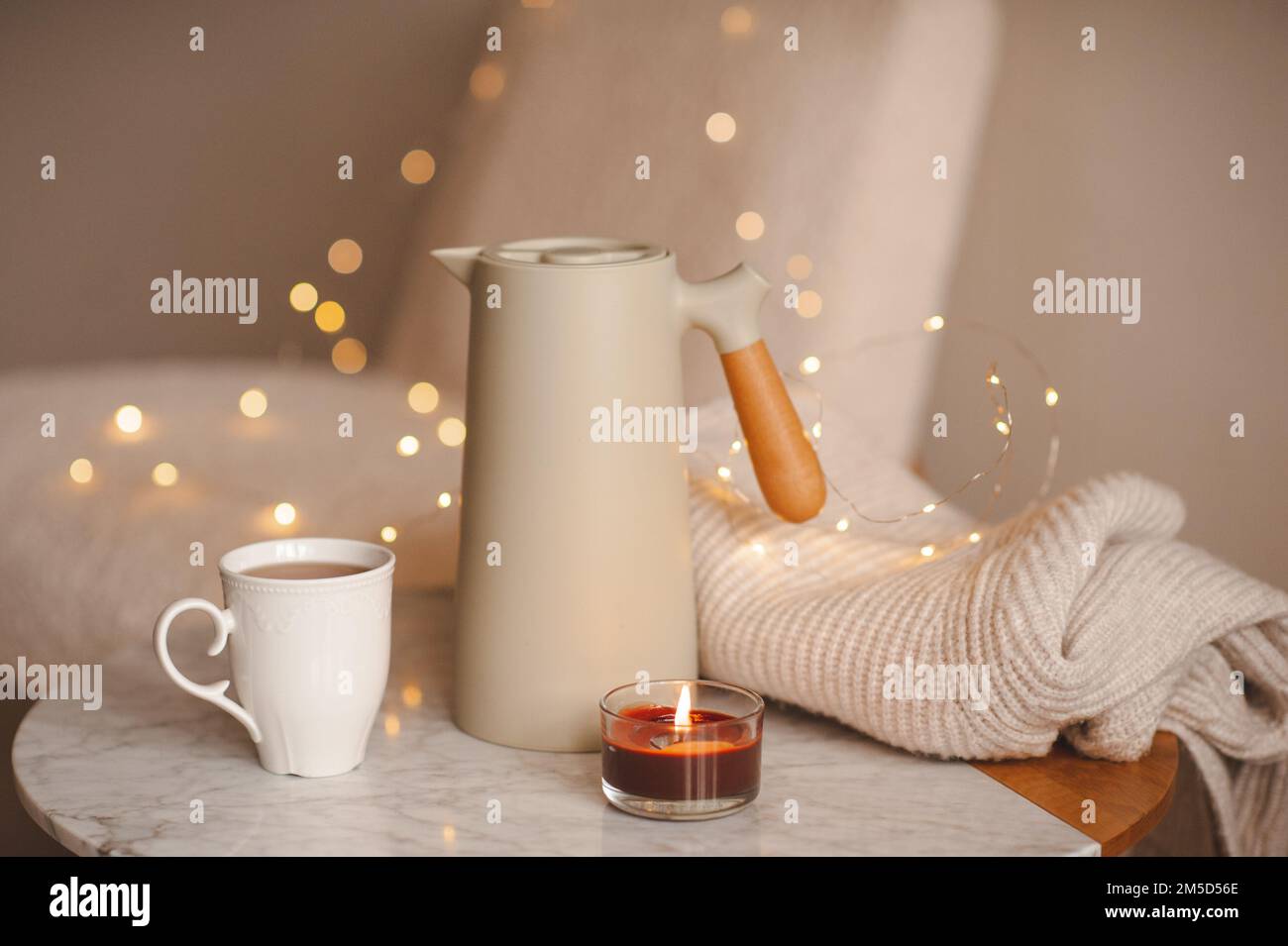 Caffettiera con una tazza di candela profumata che brucia il tè sul tavolo di marmo sopra le luci di Natale in camera da letto. Atmosfera accogliente casa. Foto Stock