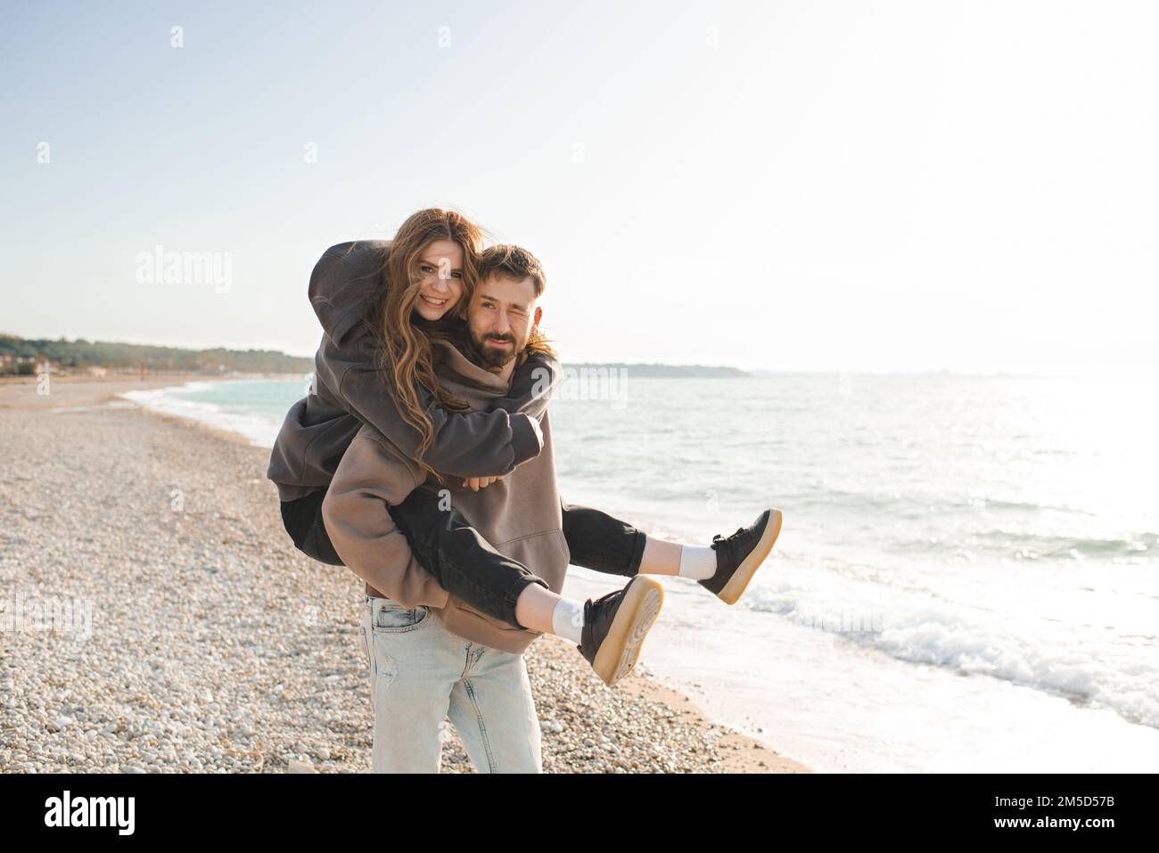 Felice coppia di giovani adulti che si divertono in spiaggia su sfondo mare insieme. Un uomo sorridente porta una bella donna sull'oceano. Relazioni. Foto Stock