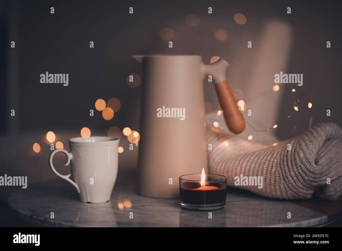 Caffettiera con una tazza di candela profumata che brucia il tè sul tavolo di marmo sopra le luci di Natale in camera da letto. Atmosfera accogliente casa. Foto Stock