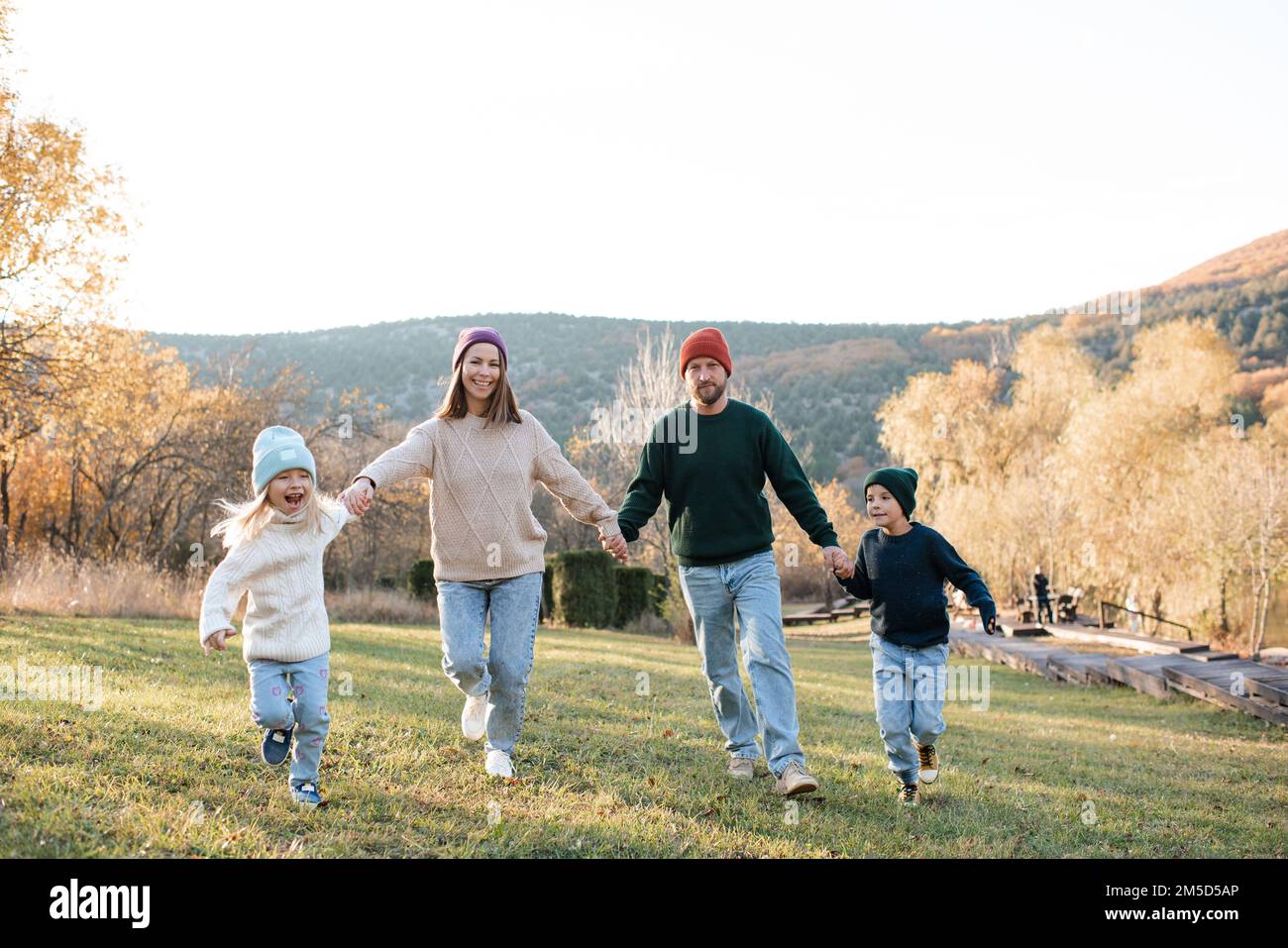 Famiglia felice che corre in scena rustica con erba verde e sfondo autunno natura insieme tenendo le mani. Allegro e felice. Foto Stock