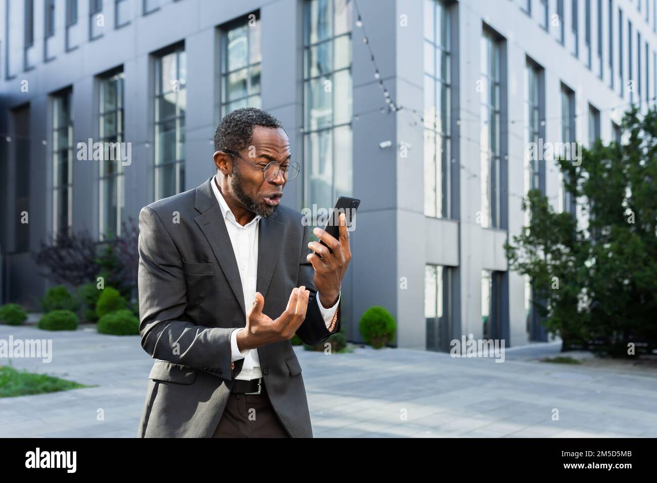 Arrabbiato capo afroamericano litigare e gridare mentre si parla al telefono, uomo anziano in tuta aziendale fuori sede ufficio. Foto Stock
