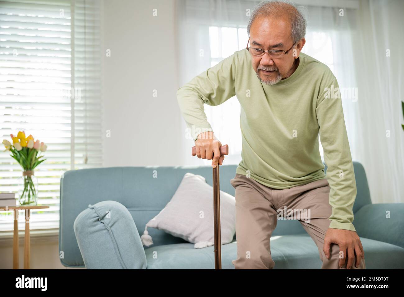 Asian Old Man con occhiali che battono per alzarsi dal divano con bastone da passeggio Foto Stock