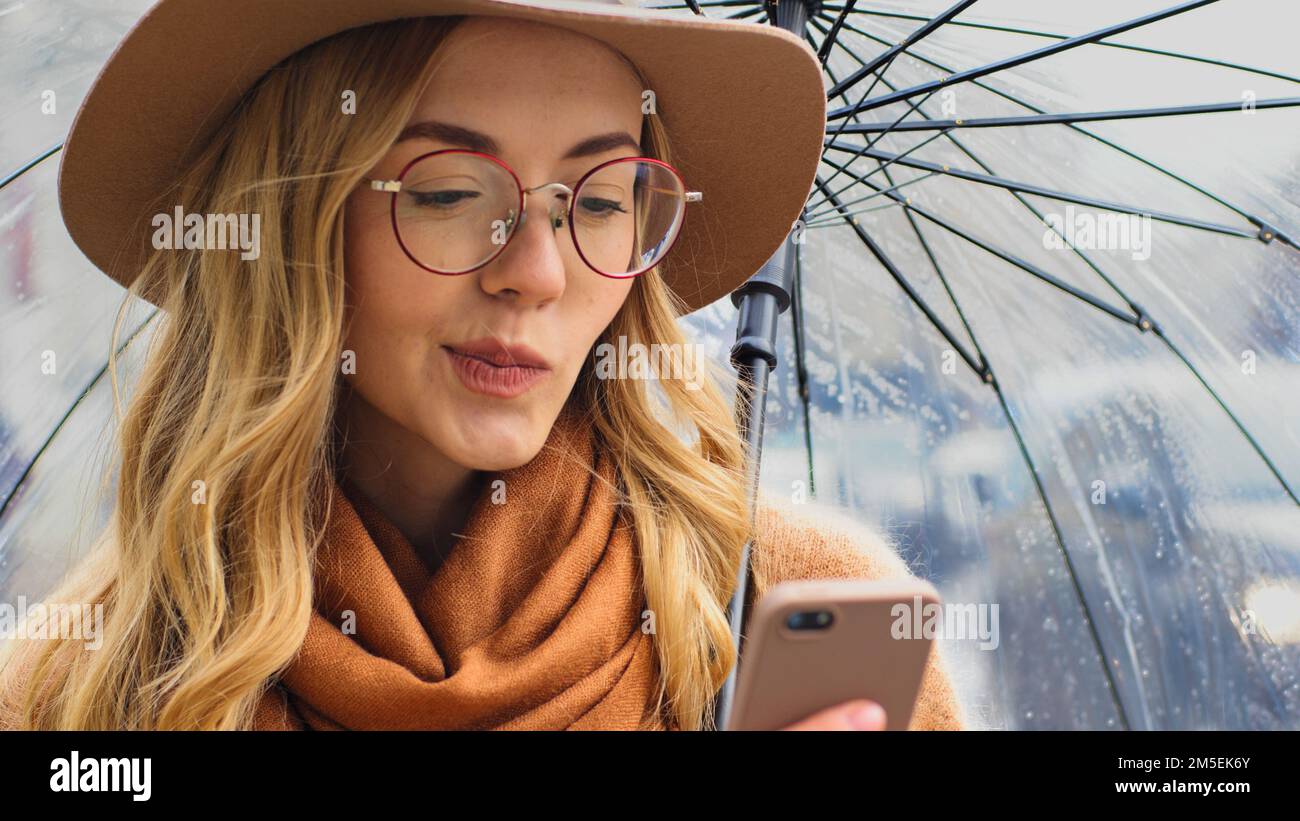 Ritratto di pensiero pensivo perplesso confuso giovane studentessa con telefono stand in strada con ombrello donna caucasica in cappello e occhiali in città Foto Stock
