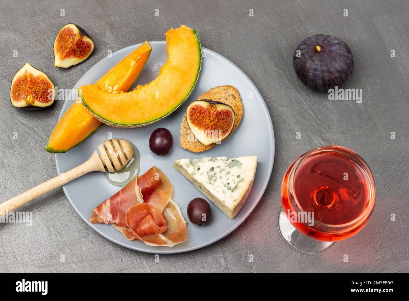 Bastone al miele e formaggio, olive brune, jamon e melone su piatto grigio. Bicchiere di vino e metà di fichi a tavola. Giacitura piatta. Sfondo grigio. Foto Stock