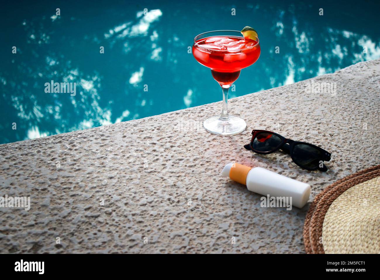 Rinfrescante cocktail rosso in piscina con crema solare, occhiali da sole e cappello di paglia Foto Stock