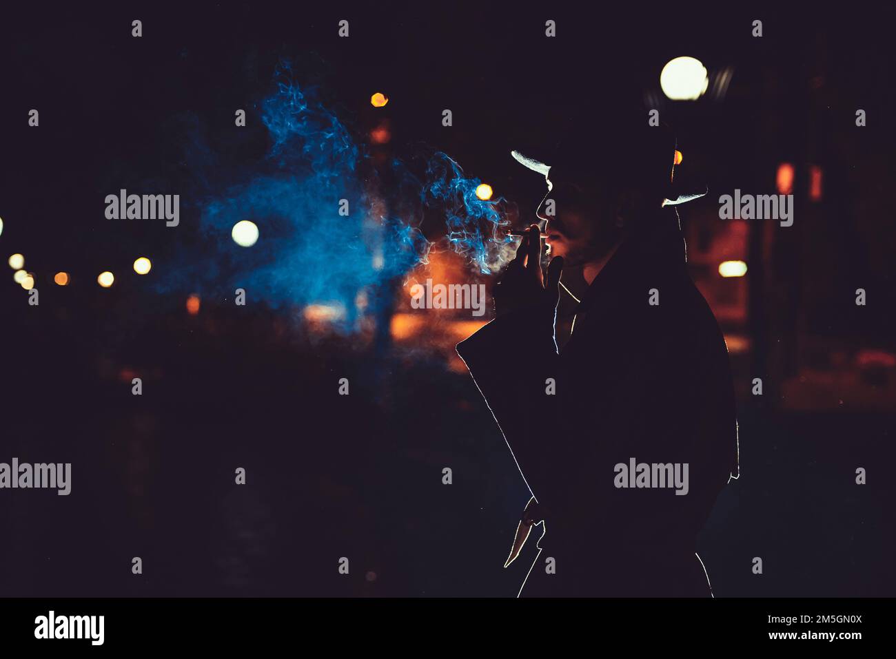 l'agente detective dell'uomo in un cappello e un impermeabile fuma una sigaretta di notte in una città piovosa nello stile del film noir Foto Stock