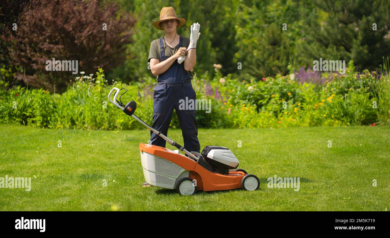 Un giovane uomo in un cappello di paglia sta mettendo i guanti per mowe un prato con un rasaerba nel suo bel giardino estivo verde floreale. Foto Stock