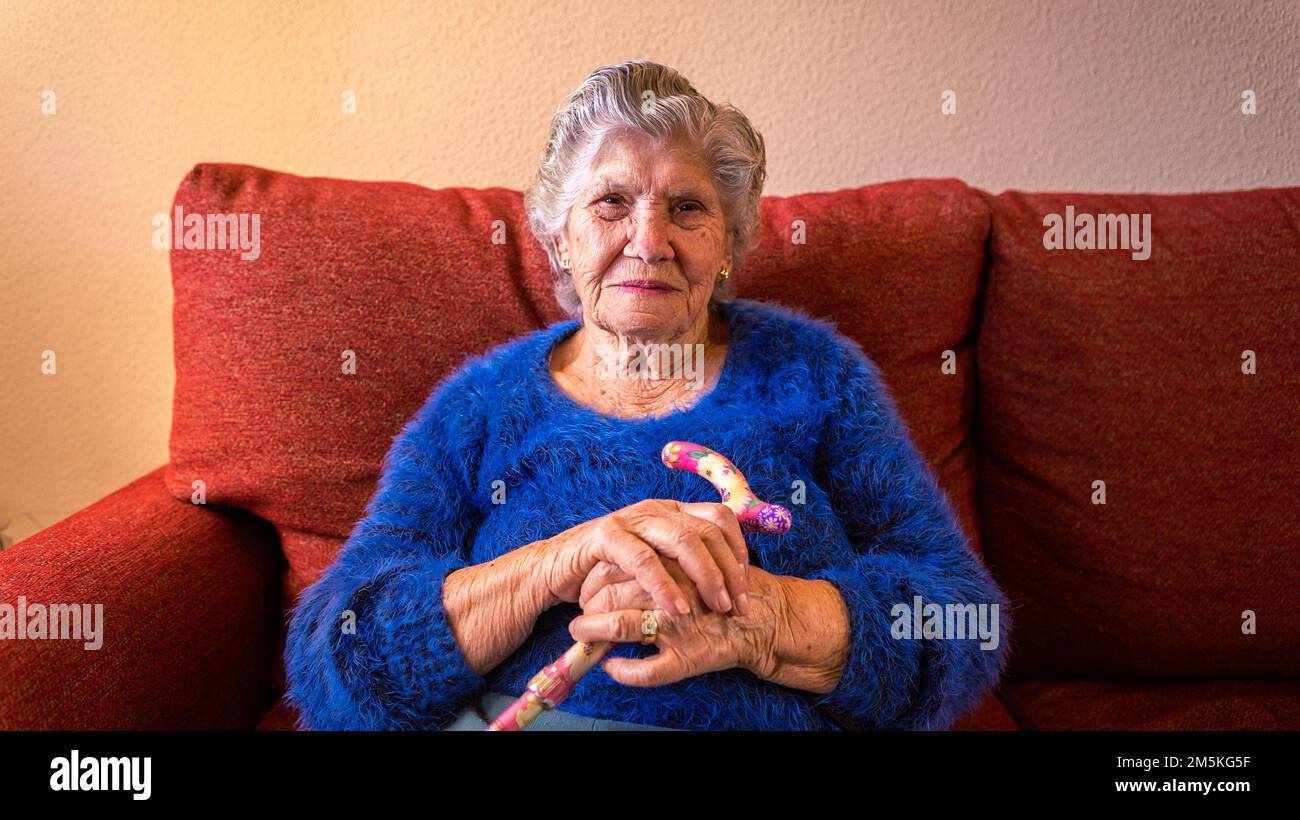Nonna che tiene un bastone mentre lei seduta sul divano in soggiorno. Donna di 90s anni sul divano a casa. Concetto di persone anziane. Foto Stock