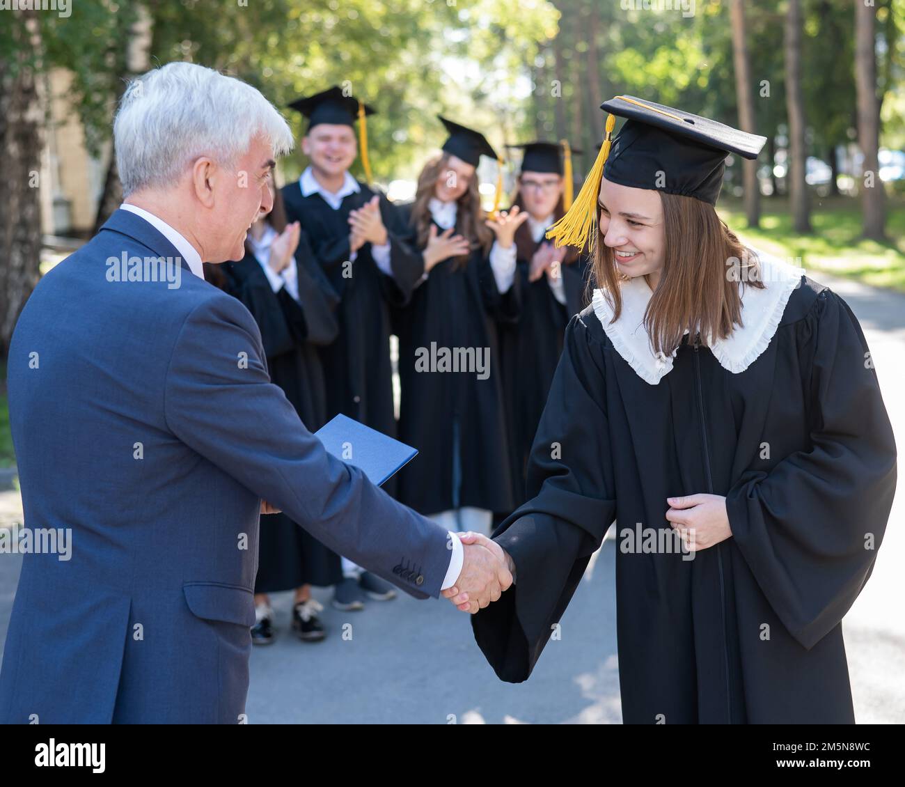 L'insegnante scuote le mani con lo studente e presenta il diploma all'aperto. Un gruppo di laureati. Foto Stock