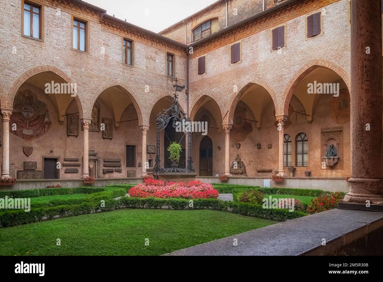 Il Chiostro della Magnolia nella Basilica di Sant'Antonio, Padova, presenta archi in mattoni gotici, pozzo in ferro battuto, giardino ben curato e luce calma. Foto Stock
