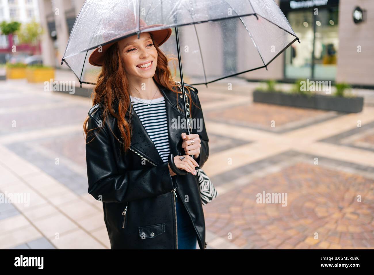 Ritratto di giovane donna allegra rossa che indossa un cappello di moda in  piedi con ombrello trasparente sulla strada della città vicino al centro in  caso di pioggia Foto stock - Alamy