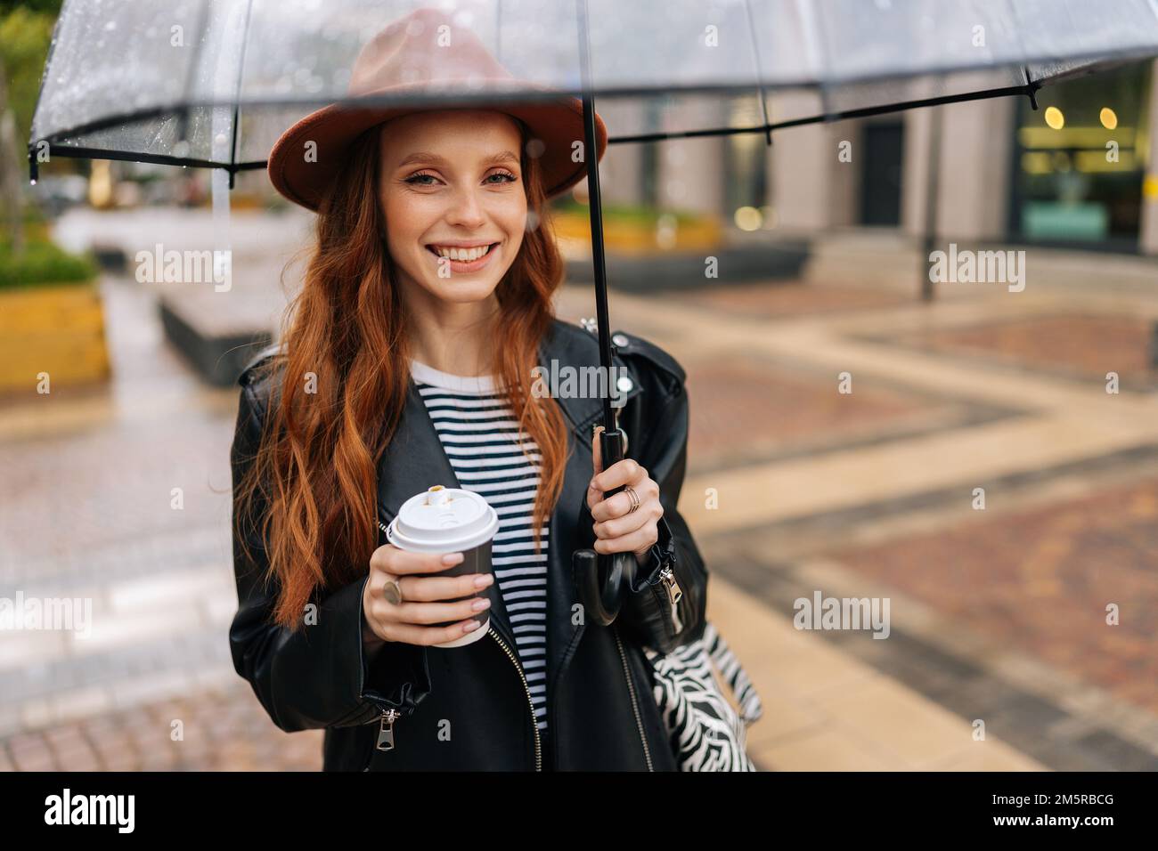 Ritratto di zenzero positivo giovane donna in elegante cappello che tiene  in mano la tazza con caffè caldo in piedi con ombrello trasparente nella  fredda pioggia autunnale Foto stock - Alamy