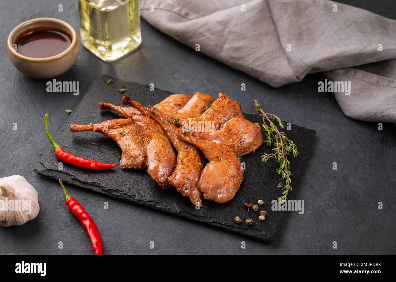 Cosce di quaglia grigliate alla griglia in marinata e spezie su ardesia nera con erbe fresche, olio e salsa su sfondo scuro. Alimentazione sana. Spazio di copia. Foto Stock