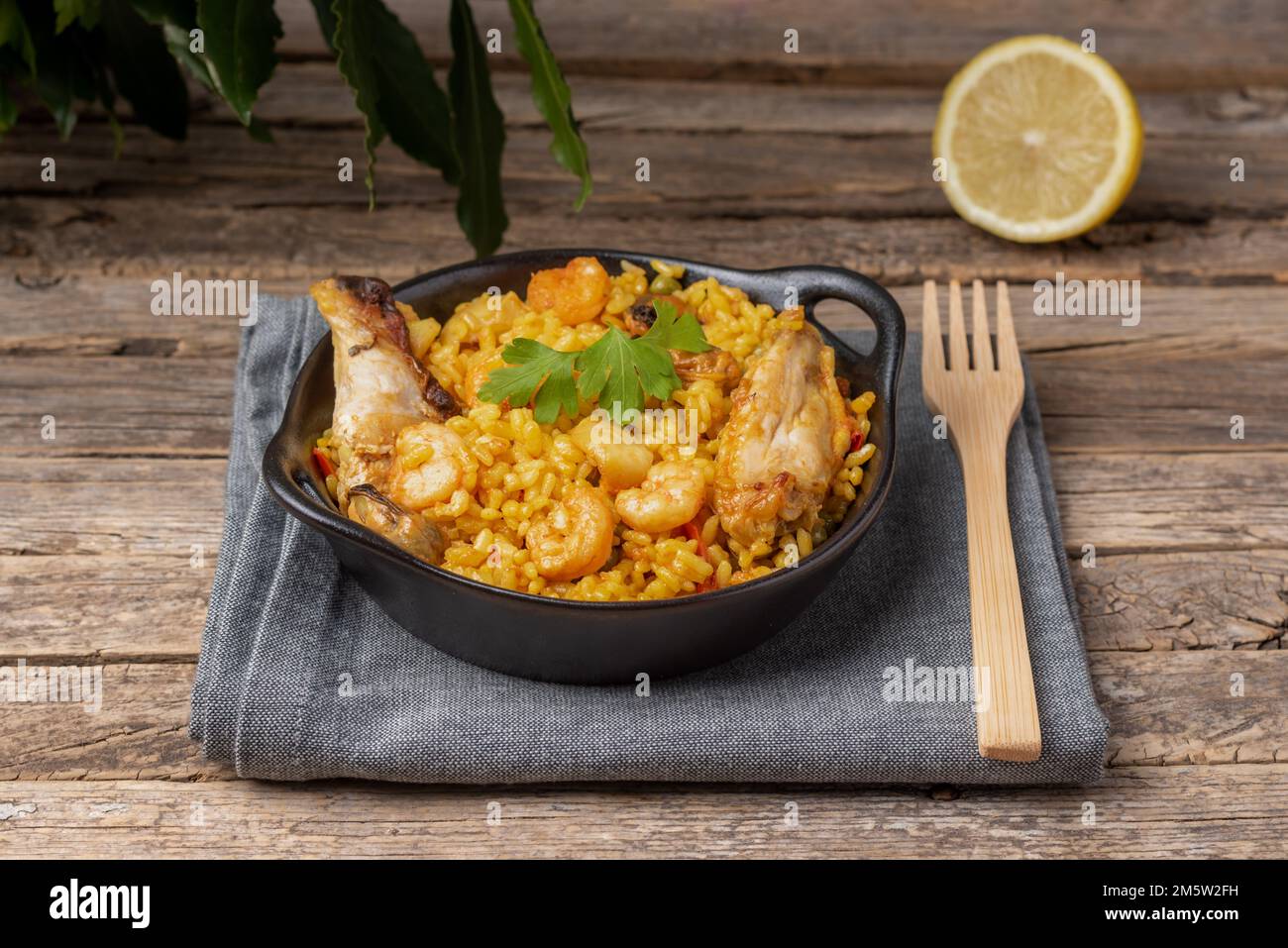 Piatto nero con paella mista, carne e pesce, su un panno da cucina grigio, una forchetta di legno e mezzo limone, su un tavolo rustico di legno. Foto Stock