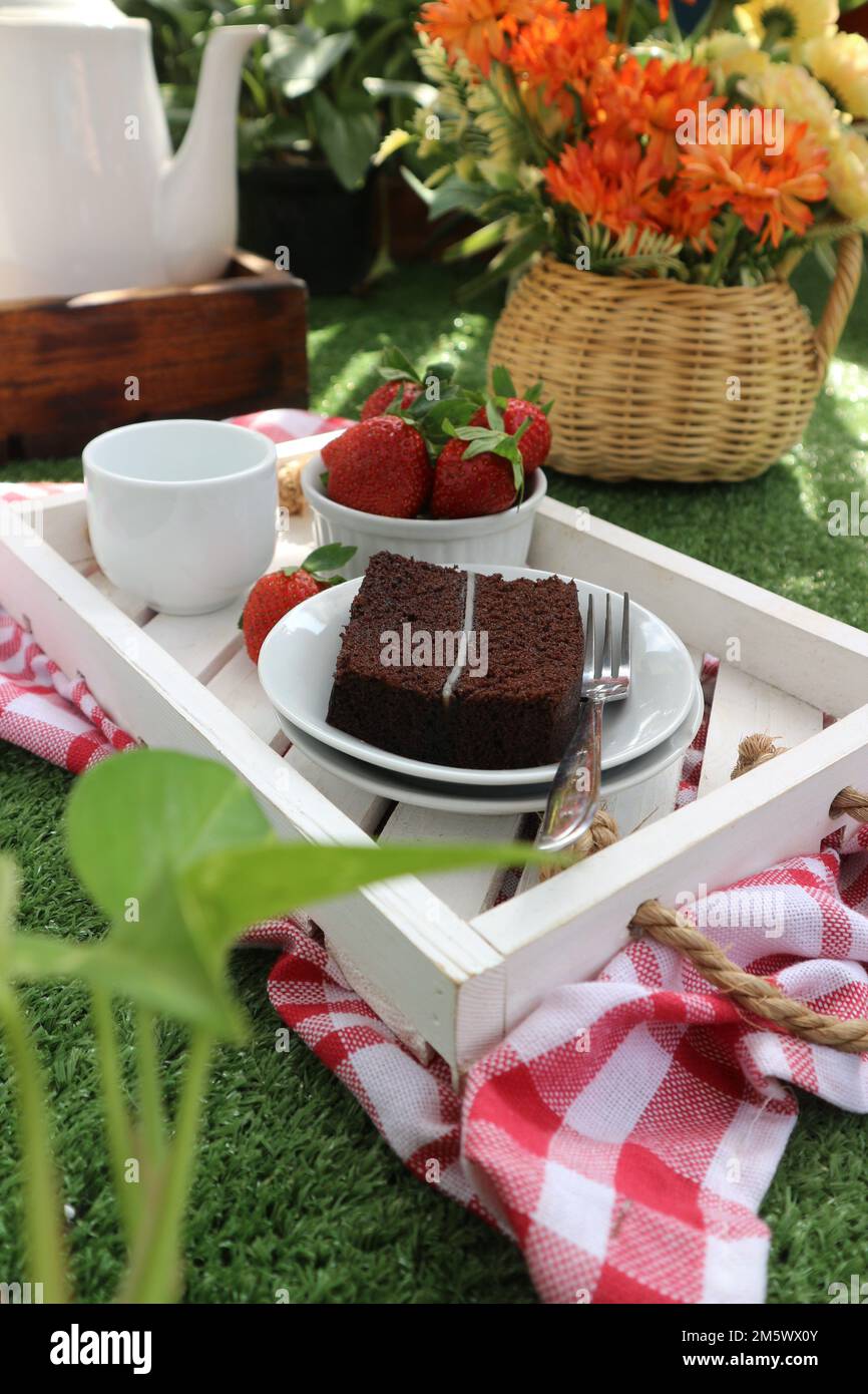 un vassoio contenente un pezzo di torta al cioccolato su un piatto bianco, una tazza di tè e una ciotola di fragole su uno sfondo di fiori in un vaso di rattan Foto Stock
