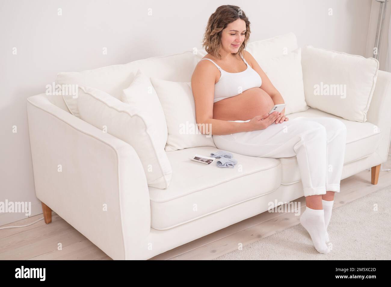 Una donna incinta siede su un divano bianco e tiene uno smartphone. Screening fetale a ultrasuoni. Foto Stock
