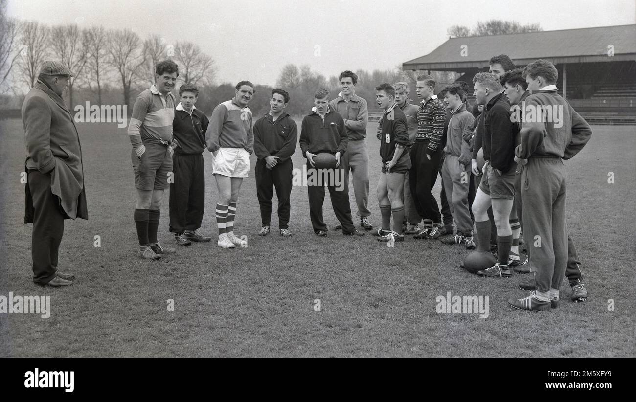 Anni '1950, storici, giovani membri della squadra di rugby delle English Schools in piedi fuori con il loro allenatore, in un impermeabile e cappello piatto, prima dell'inizio di una sessione di allenamento, Inghilterra, Regno Unito. Divisa sportiva del giorno in mostra, con top in cotone e tute sportive. Foto Stock