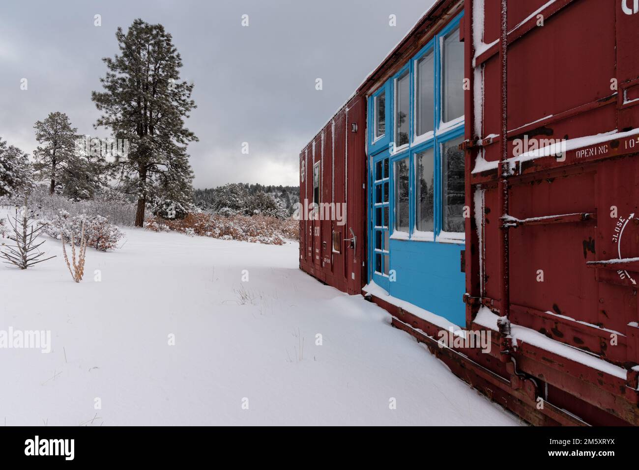 Treno rosso scuro con grandi finestre e porte incorniciate da porte e finestre color turchese in un paesaggio innevato nel New Mexico settentrionale, Stati Uniti. Foto Stock