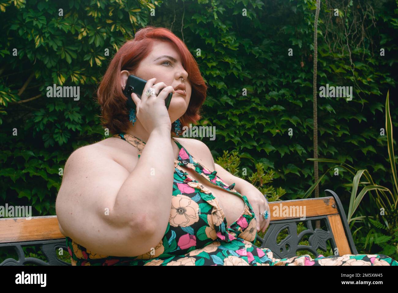 giovane donna caucasica latina di etnia argentina, più modello taglia, capelli corti di colore rosso, seduta rilassata rispondendo a una chiamata telefonica in giardino, Foto Stock