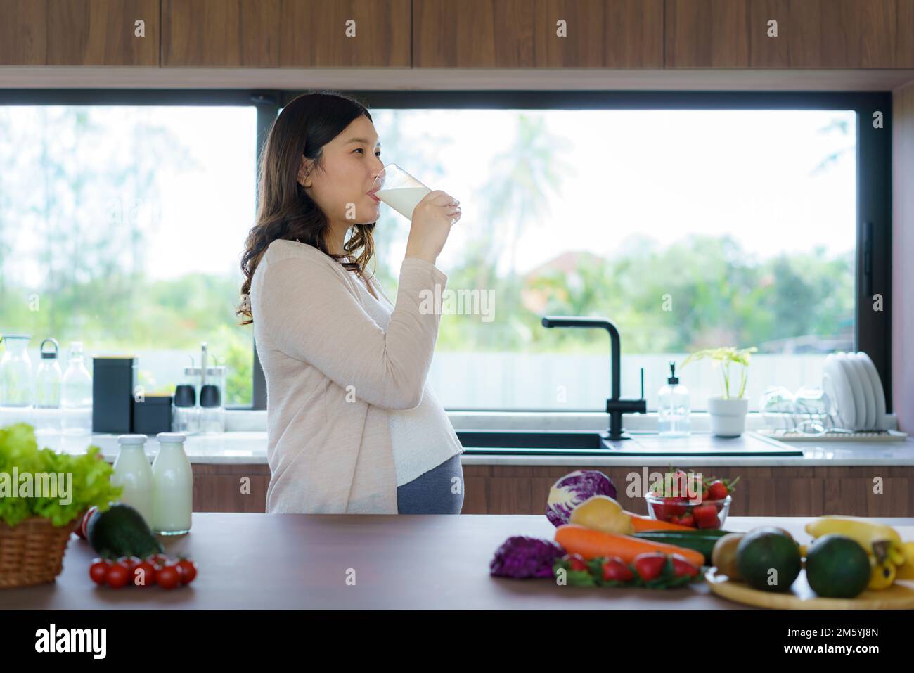Affascinante donna asiatica incinta che beve latte dal vetro. Donna attraente ha bisogno di proteine e calcio per il suo bambino e rende la sua sana e forte. Foto Stock