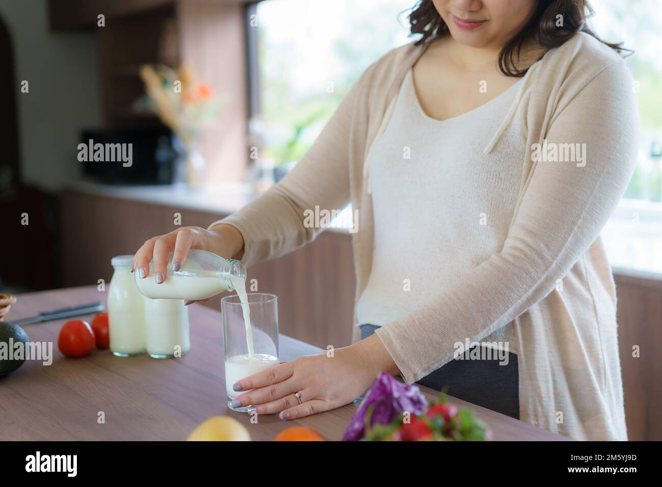 Affascinante donna asiatica incinta sta tenendo una bottiglia di latte e versando il latte in un bicchiere. Donna attraente bisogno di proteine e calcio per il suo bambino e mak Foto Stock