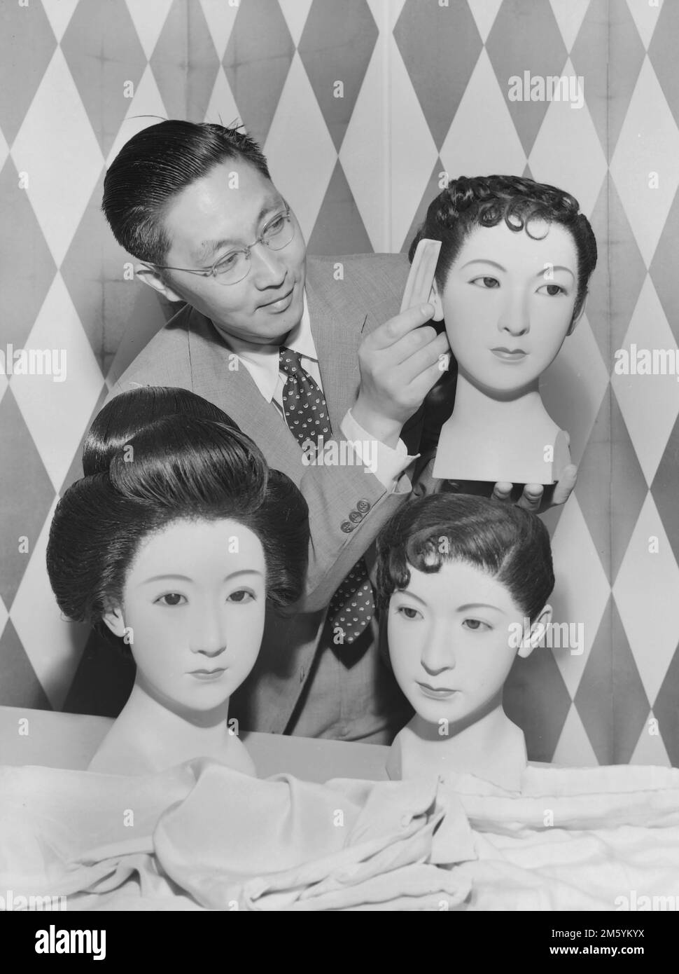 Un hairstylist americano asiatico funziona sui hairpieces sui mannequins americani asiatici, ca. 1960. Foto Stock