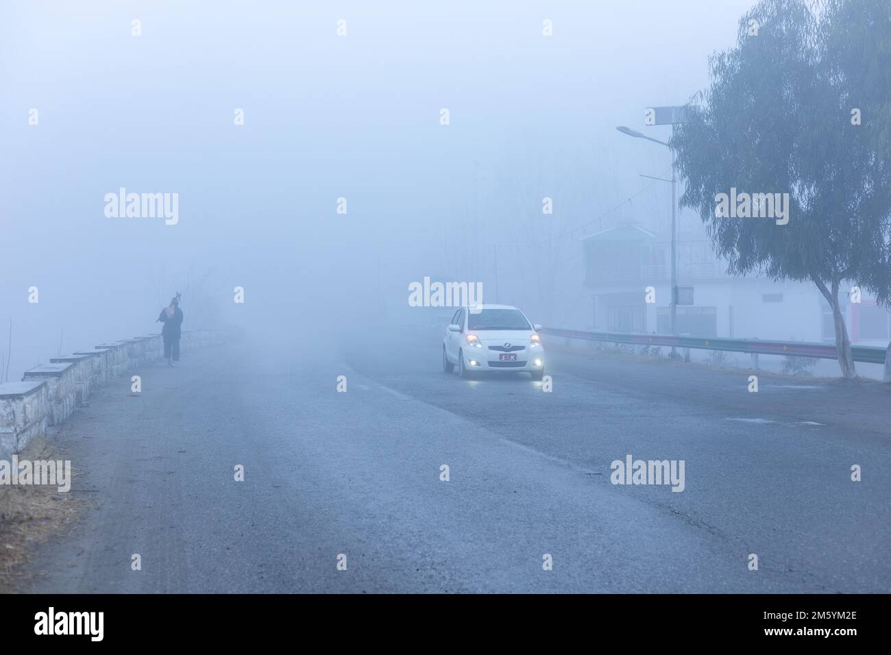 Uomo che cammina in nebbia fitta la mattina presto sul lato della strada con auto che arriva su una strada con scarsa visibilità Foto Stock