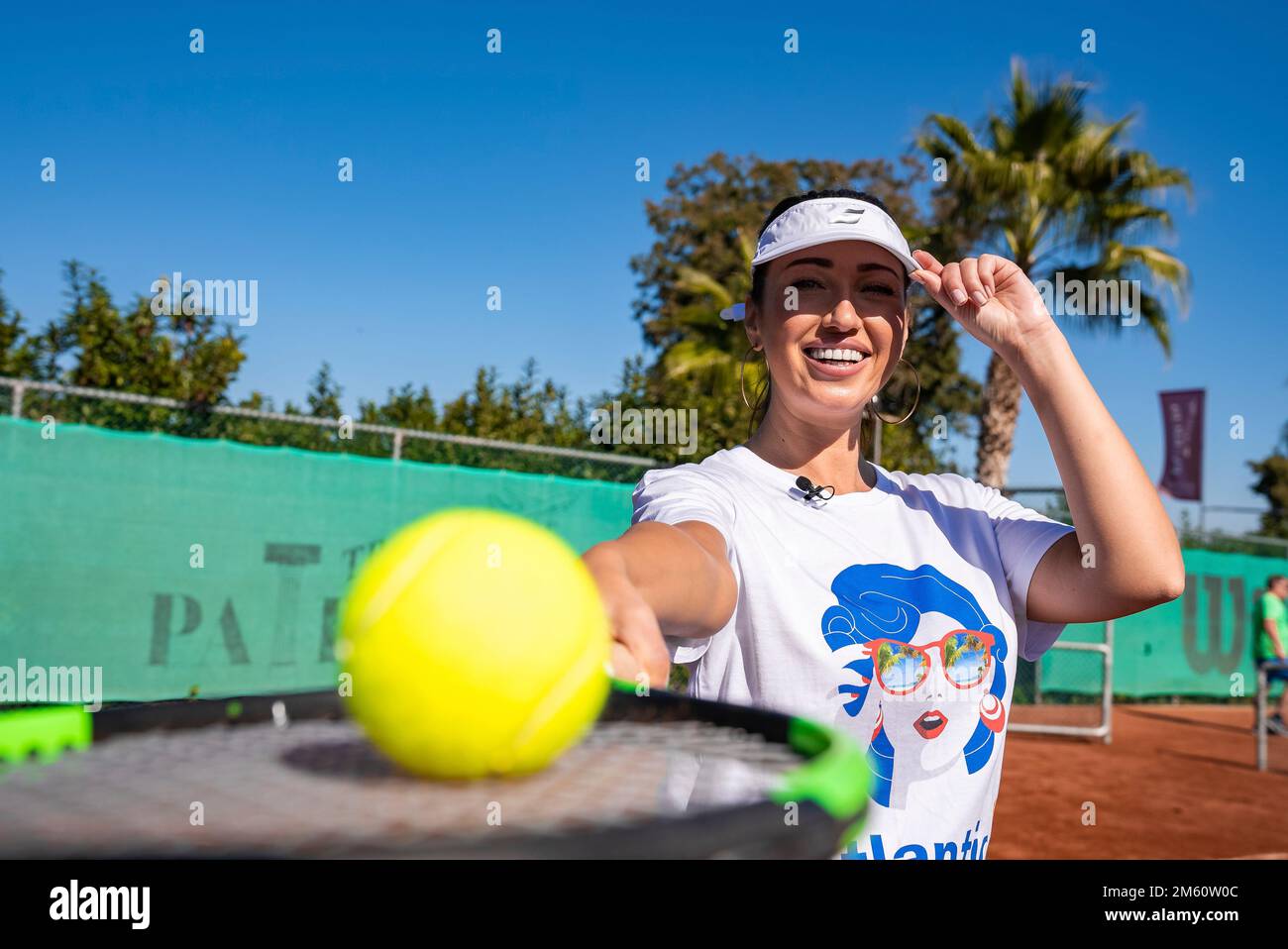 Donna felice che indossa il cappuccio della visiera che tiene la racchetta da tennis e la palla in campo Foto Stock