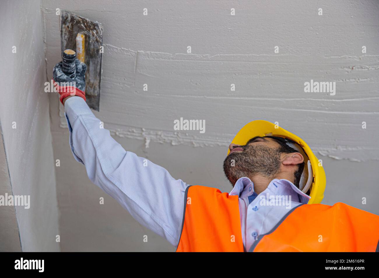 Un intonaco in costruzione indossa tute e un cappello. Sta intonacando un muro con stucco di finitura utilizzando un coltello stucco. Lavori di riparazione e costruzione c Foto Stock