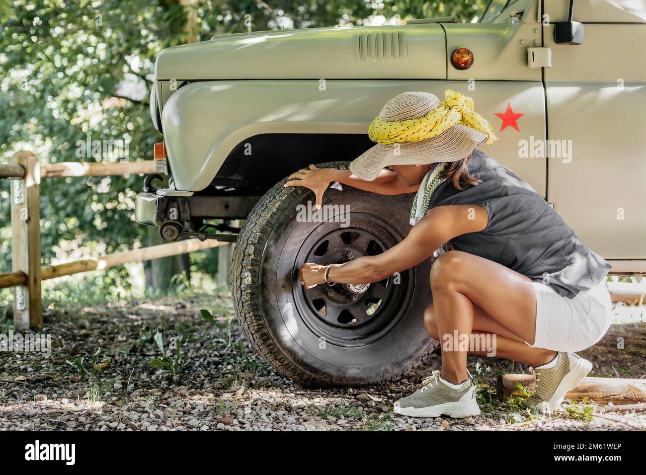 vista del profilo di una donna avventurosa che indossa un cappello e una sciarpa, controllando le condizioni degli pneumatici del suo veicolo fuoristrada durante una pausa. Foto Stock