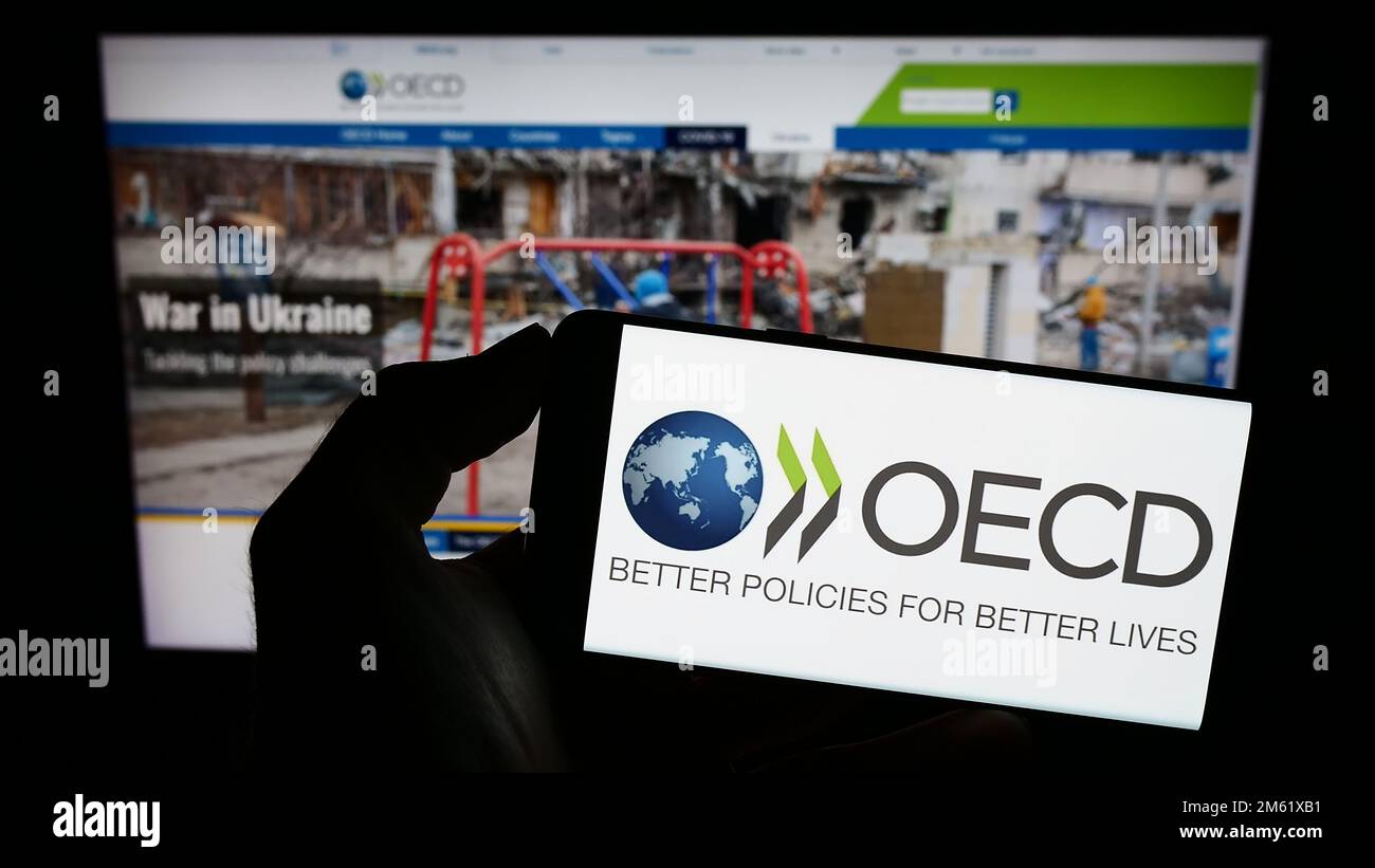 Persona che tiene il cellulare con il logo dell'organizzazione intergovernativa OCSE sullo schermo di fronte alla pagina web. Messa a fuoco sul display del telefono. Foto Stock